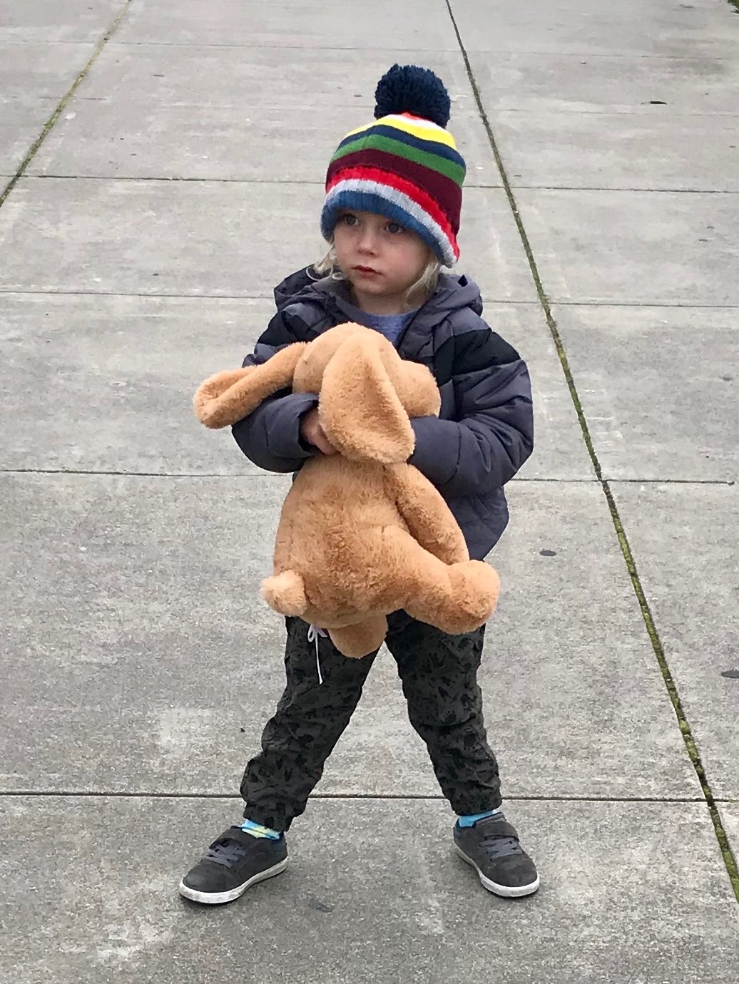 Toddler Elijah on a sidewalk wearing a striped pom-pom hat and winter coat, holding a stuffed dog