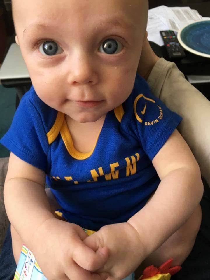 Baby Elijah in a Golden State Warriors Kevin Durant shirt, looking at the camera with big blue eyes